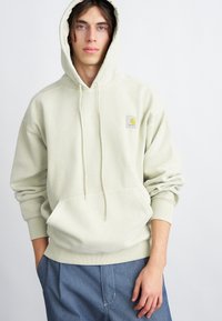 Sudadera verde claro hecha de un tejido suave y texturizado, con un bolsillo frontal y capucha con cordón. Presenta un pequeño logotipo de la marca en el pecho.