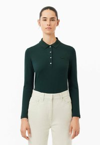Mörkgrön långärmad polo tröja med knapplist och broderad logotyp, ihop med ljusa högmidjade byxor.