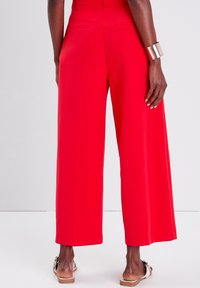 Pantalon rouge à jambes larges avec une texture lisse, taille haute et poches arrière. Accessoires : un bracelet manchette doré et des sandales décontractées.