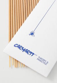L'emballage d'encens Carhartt WIP présente une carte blanche avec du texte bleu et un graphique d'araignée, accompagnée de plusieurs bâtons d'encens fins de couleur brun clair.