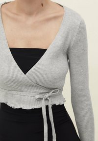 Top avvolto grigio a coste con maniche lunghe, scollatura a V e orlo arricciato; legato in vita con un nastro in tessuto coordinato. Sottostrato di colore nero.