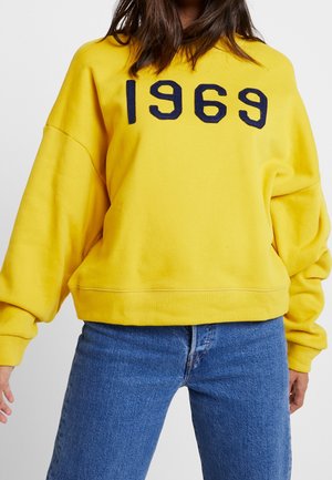 Sweatshirt jaune avec broderie marine "1969", coupe décontractée et ourlet côtelé. Associé à un jean bleu clair. Material en coton doux.