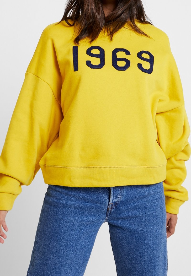 Felpa gialla con ricamo blu navy "1969", vestibilità rilassata e bordo a costine. Abbinata a jeans azzurri chiari. Materiale in cotone morbido.