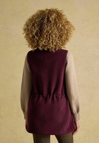 Burgunderfarbene ärmellose Weste mit einer taillierten Detail, kombiniert mit einem hellbeigen Langarmshirt, das strukturierte gestrickte Ärmel aufweist.