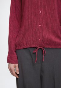 Bordeauxrode lange mouw shirt met een gestructureerd patroon, knoopsluiting aan de voorkant en een trekkoord in de zoom, gecombineerd met grijze broek. Zachte stof met een ontspannen pasvorm.