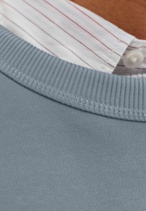 Grå bomullströja med ribbad krage, lager på lager över en vit skjorta med ljusröda vertikala ränder.