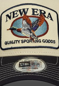 Gorra crema con visera oscura que presenta un diseño bordado de un pato y texto. Etiqueta de tamaño ajustable en la parte interior. Tela duradera.