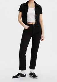 Sort struktureret kortærmet skjorte over en hvid cropped tank top, parret med højtaljede sorte jeans og sorte sneakers med hvide accenter.