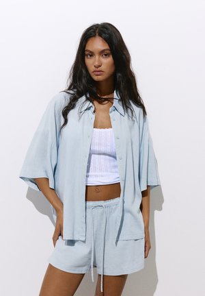 Femme aux longs cheveux foncés porte une chemise bleu clair oversize boutonnée et un short assorti par-dessus un débardeur court blanc, debout devant un fond blanc.