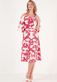Off-the-shoulder white dress with bold red floral patterns, tiered design, flared sleeves, and a fitted waist, paired with white heels.