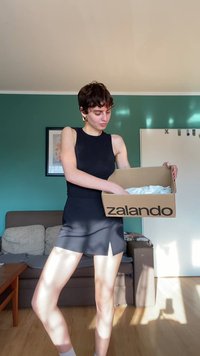Vestito nero senza maniche con corpetto aderente e orlo leggermente svasato, abbinato a una scatola di cartone Zalando contenente tessuto bianco.