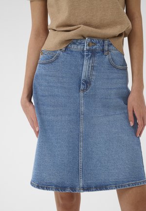 Kvinde iført en knælængde blå denim nederdel med frontsyning og lommer, kombineret med en beige top.