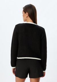 Svart stickad cardigan med kortad skärning, som har en vit ribbad kant längs halsringningen och fållen. Långa ärmar med textilstruktur.
