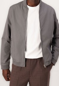 Šedá bomber bunda se zipem, manžetami a dvěma bočními kapsami, oblečená přes bílé tričko a hnědé kalhoty s vzorem.