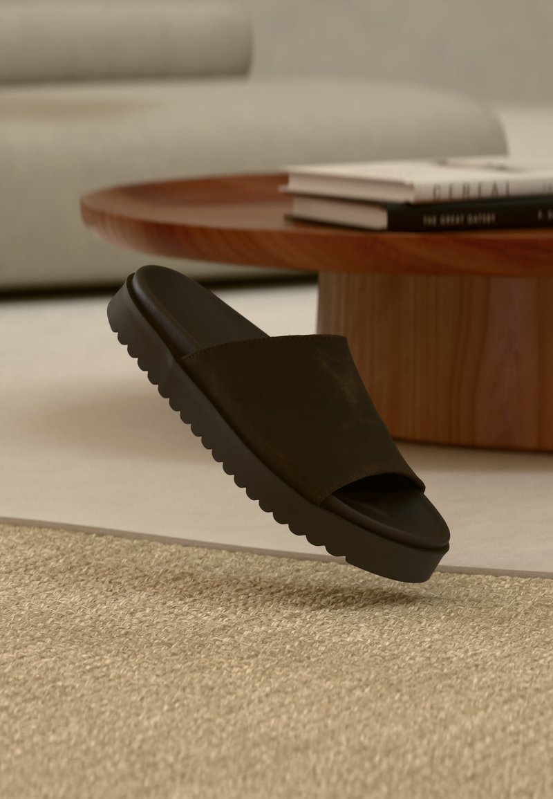 Sandale à glissière noire avec semelle épaisse flottant au-dessus d'un tapis beige texturé dans un salon moderne près d'une table basse en bois avec des livres.