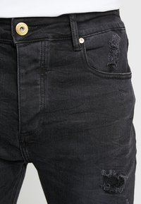 Vaqueros de denim negro con un diseño clásico que presenta un desgaste visible en el bolsillo delantero y un cierre con botón dorado.