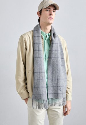 Grå uldscarf med et blåt rutenet mønster og frynser, båret over en lysegrøn skjorte, komplementeret af en beige jakke og hat.