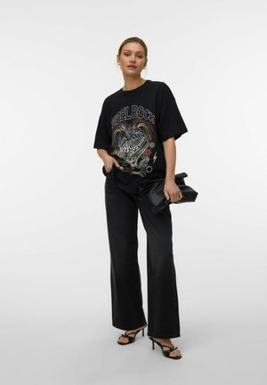 Femme portant un t-shirt noir graphique "REBEL ROCK", pantalon noir à jambes larges, sandales à talons noires, tenant une pochette noire sur fond blanc.