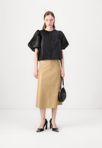 Schwarzes Satin-Oberteil mit Puffärmeln, kombiniert mit einem beigen Bleistiftrock. Schwarze High-Heels und eine kleine schwarze Handtasche vervollständigen den Look.