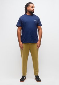 Άνδρας με πλεξούδες, φορώντας μπλε ναυτικό t-shirt με τσέπες, χακί παντελόνι jogger και μαύρα αθλητικά παπούτσια, στέκεται μπροστά σε απλό λευκό φόντο.