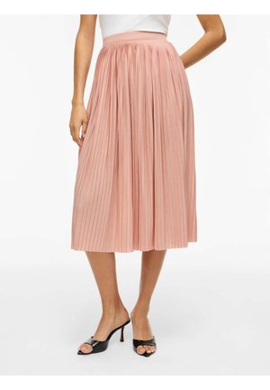 Kobieta w wysokim, plisowanym różowym spódnicy midi w połączeniu z czarnymi sandałami na obcasie z odkrytymi palcami na białym tle.