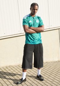 Ung man med flätat hår, klädd i en grön sporttröja, svarta vida shorts, vita strumpor och svarta skor, stående med armarna i kors utomhus.