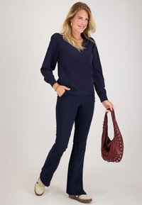 Marineblauwe blouse met lange mouwen en een V-hals, gecombineerd met marineblauwe flared broeken. Houdt een bordeaux handtas met zilveren accenten vast; neutrale sneakers.