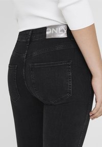 Jeans de mezclilla negra con un diseño de cintura alta, dos bolsillos traseros y un parche de logo plateado en la cinturilla. Textura suave, forma ajustada.