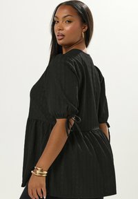 Donna con lunghi capelli scuri che indossa una blusa nera a texture con maniche corte legate e orecchini e bracciali dorati, mentre guarda oltre la spalla.
