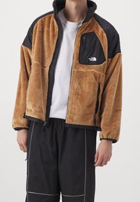 Giacca in pile marrone con accenti in nylon nero, colletto alto, tasca con zip, polsini elasticizzati e tasche laterali; indossata sopra una camicia bianca.