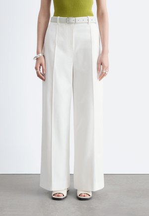 Femme portant un pantalon blanc taille haute à jambes larges avec une ceinture blanche, un haut sans manches vert, un bracelet et des bagues argentés, ainsi que des chaussures blanches ouvertes aux orteils.