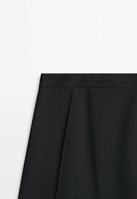 Section d'une jupe plissée noire montrant la ceinture et la texture du tissu sur fond blanc.