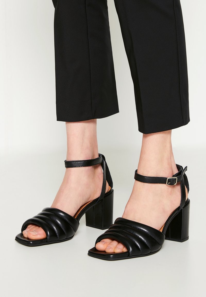 Mustat korkokengät sandaalit, joissa on neliön muotoinen varvas ja nilkkahihna. Valmistettu sileästä nahasta, jossa on päällä quiltattu kuvio ja tukeva lohkare korko.