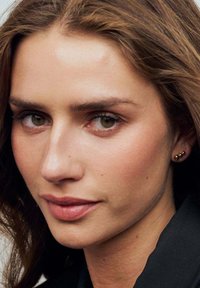 Närbild av ett ansikte med medelhårig brun hårfärg, naturlig makeup, gröna ögon och guldörhängen. Slät hud med subtila fräknar. Svart topp.