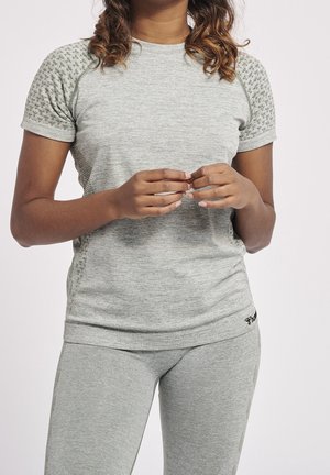 Persoon met lichtgrijs kort mouw shirt en bijpassende broek, handen voor zich gevouwen, met krullend bruin haar zichtbaar.