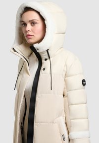 Krémová péřová bunda s kapucí lemovanou kožešinou, polstrovaný design, černý zip a akcent s logem, s předním zipem a šňůrkami.