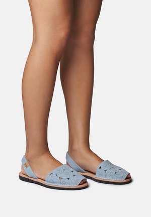 Noge v svetlo modrih odprtih slingback sandalih z zapletenimi izrezanimi vzorci in črnimi ravnimi podplati na belem ozadju.