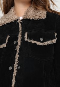 Svart mockajacka med en fluffig brun krage, två bröstfickor med lock och tryckknappar. Mjuk textur med kontrasterande detaljer.