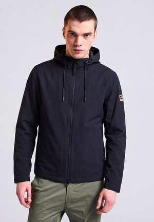 Junger Mann trägt eine schwarze Kapuzenjacke, die zugezippt ist, und steht vor einem schlichten, hellen Hintergrund, die Hände entspannt seitlich.