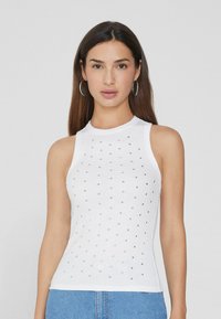 Top de tirantes blanco con relieve que presenta accentos cuadrados plateados en la parte delantera y un cuello alto. La tela parece suave y ajustada.
