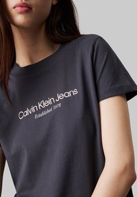 Camiseta de algodón gris oscuro con la leyenda "Calvin Klein Jeans" y "Fundada en 1978" en tinta rosa claro impresa en la parte delantera. Mangas cortas.