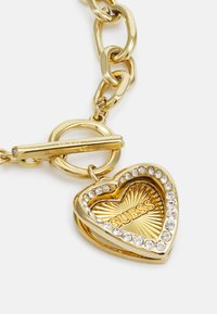 Collier à maillons en or avec un pendentif en forme de cœur gravé du mot "GUESS", entouré de strass et d'un intérieur texturé en rayons de soleil.