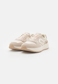 Beige athletic shoes with a mix of suede and textile materials. Features a lace-up design, textured panels, and a rubber sole.