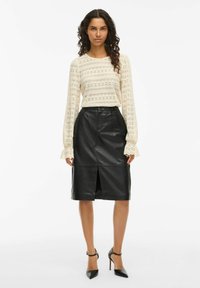 Vrouw met krullend haar, gekleed in een crèmekleurige kanten blouse met lange mouwen, een zwarte leren rok tot de knie met een voorkrui en zwarte hoge hakken.