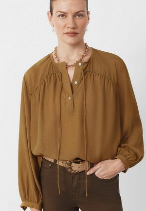 Kvinde iført en sennepsgul bluse med knapper og lange ærmer, stoppet ned i mørke bukser, accessoriseret med en kædehalskæde og bælte med bladspænde.