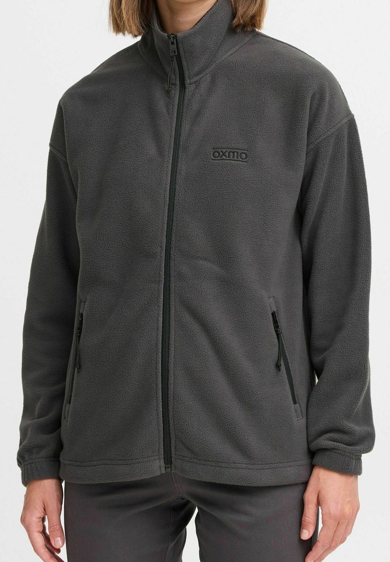 Grå fleecetrøje med høj hals, lynlås foran og sidelommer. Har et diskret logo på brystet og blødt, tekstureret materiale.