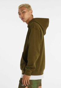 Felpa verde oliva in tessuto morbido con tasca a marsupio, cappuccio con coulisse regolabile e polsini a costine. Indossata sopra una camicia bianca.