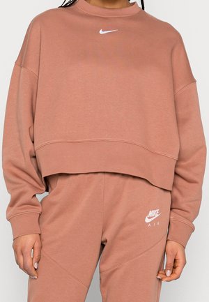 Personne portant un ensemble sweat-shirt et pantalon de survêtement Nike Air rose poussiéreux assorti avec des logos Nike blancs sur la poitrine et la cuisse.