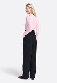 Langarmige roze top met een gerimpelde kraag en een keyhole achterkant, gecombineerd met hooggesneden, wijde zwarte broeken. Schoenen zijn bordeauxrood.