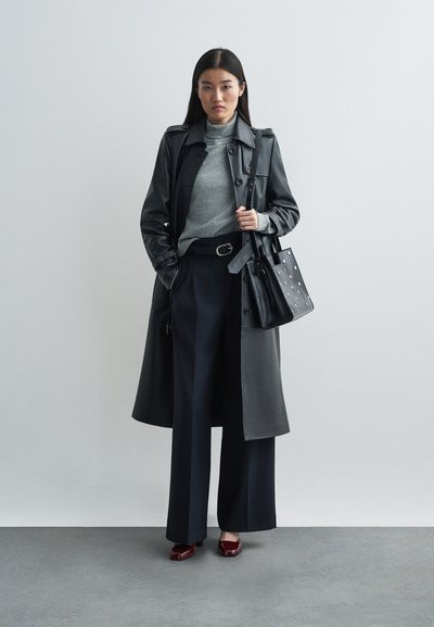 Zwarte leren trenchcoat over een grijze coltrui, gecombineerd met marineblauwe wijde broek. Accessoires zijn een zwarte tas met witte stippen en rode schoenen.